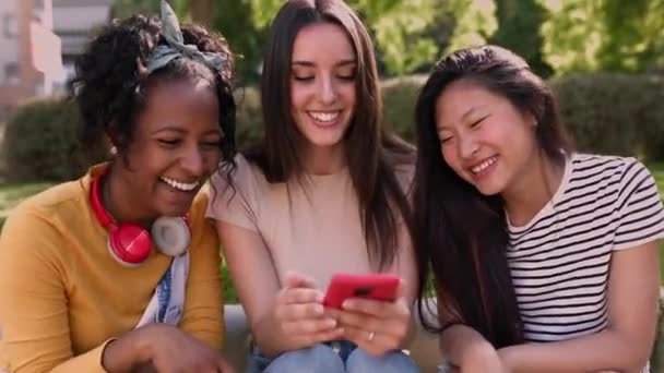 ハッピーな若い多人種の女性の友人は 屋外で一緒にスマートな携帯電話を使用しています 外で夏を過ごしながらスマートフォンを保持する3千年もの多様な女性 — ストック動画