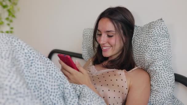 若いミレニアル世代の少女は ベッドで休んでいる携帯電話を使用して楽しんでいます 目覚めた後 スマートフォンアプリで面白いメッセージを読むミレニアム女性 — ストック動画