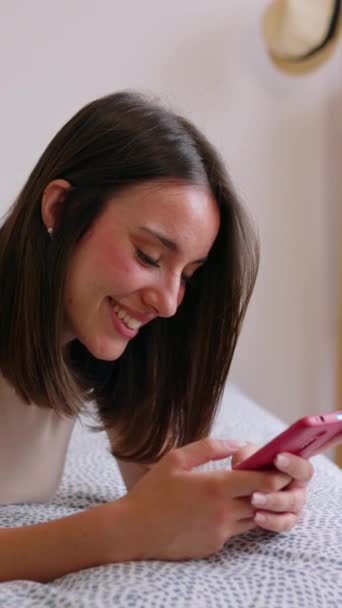 漂亮的年轻女人在家里休息的时候用手机 笑着漂亮的女孩躺在床上看着智能手机屏幕 社交媒体和技术概念 垂直屏幕视频 — 图库视频影像