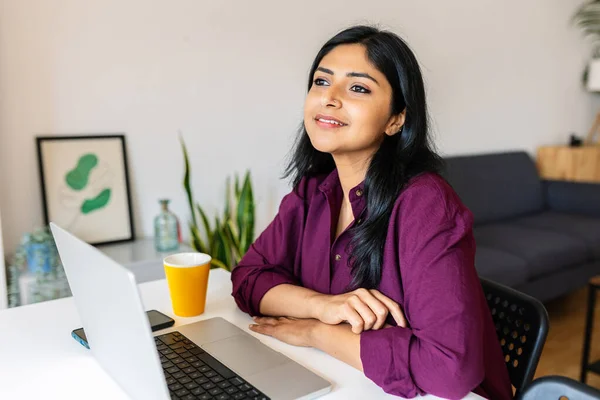 Thoughtful indian woman sitting with laptop on table while looking away at home