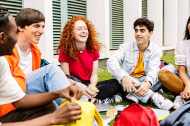 Çeşitli kolej öğrencileri gülümsüyor ve kampüs kolejinde birlikte oturup, çim parkında oturarak eğleniyorlar. Gerçek gençler ve eğitim kavramı.