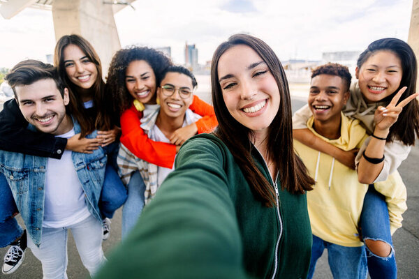 Молодая группа разнообразных студентов веселится на открытом воздухе. Женщина улыбается на камеру, делая селфи с друзьями на заднем плане. Концепция молодежного сообщества.