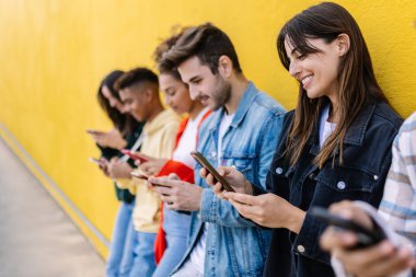 Dışarıda akıllı telefonlarını kullanan mutlu, çok ırklı bir grup genç arkadaş. Gen z insanları sosyal medya uygulamasına bağımlı, sarı arkaplan üzerinde cep telefonundaki içeriği izleyip paylaşıyorlar.