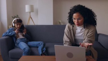 Kulaklıklı genç bir kadın diz üstü bilgisayarda çalışıyor çocuğuyla birlikte evde cep telefonu kullanıyor. Aile uzlaşması, evde çalışmak ve teknoloji yaşam tarzı kavramı.