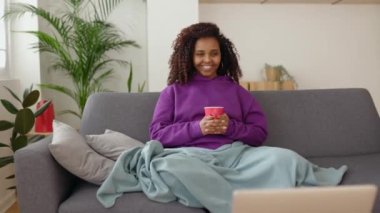 Genç ve mutlu Afrikalı bir kadın kanepede oturup dizüstü bilgisayarda film izliyor. Battaniyeyle kaplanmış Afro-Amerikalı bir kız çevrimiçi yayın platformunda filmden zevk alıyor. Hafta sonu eğlence konsepti.