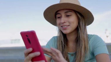 Yazlık şapkalı mutlu Latin kadın dışarıda cep telefonu kullanıyor, internette sörf yapıyor ya da sosyal ağ uygulamasında medya içeriğini izliyor. Teknoloji yaşam tarzı konsepti.