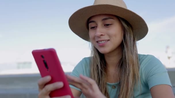 夏のハッピーな若いラテン系女性は 屋外に座って携帯電話を使用したり インターネットをサーフィンしたり ソーシャルネットワークアプリでメディアコンテンツを見たりしています 技術ライフスタイルコンセプト — ストック動画