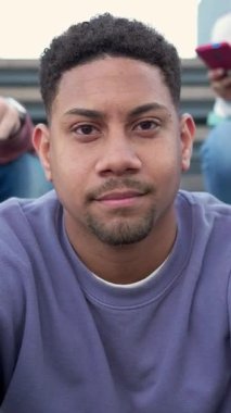 Üniversite kampüsünde kameraya gülümseyen mutlu genç Latin öğrenci arka planda sınıf arkadaşlarıyla merdivenlerde oturuyor. Dikey 9: 16 ekran görüntüsü.