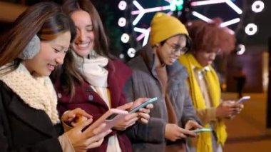 Genç mutlu arkadaşlar, kış mevsiminde şehrin sokaklarında cep telefonlarını kullanarak sıraya girip eğleniyorlar. Mobil uygulamadan sosyal medya içeriğini izleyen gençler. Ön görüntü görüntüsü.