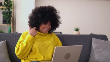 Genç, mutlu, yetişkin bir Latin kadın laptopta ailesiyle ya da arkadaşıyla konuşuyor. Neşeli bir kadın evdeki koltukta bilgisayar kullanarak çevrimiçi toplantı yapıyor. Teknoloji konsepti.