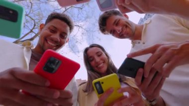 Açık havada akıllı cep telefonları kullanan genç milenyum insanlarının düşük açılı görüntüsü. Akıllı telefon ekranına bakan neşeli öğrenci arkadaşları sosyal medya içeriğinin keyfini çıkarıyorlar