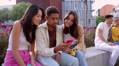 Çok ırklı bir grup genç, açık havada cep telefonu kullanırken eğleniyor. Gülümseyen öğrenci topluluğu ellerinde akıllı cep telefonları olan insanlar şehrin caddesinde birlikte oturuyorlar..