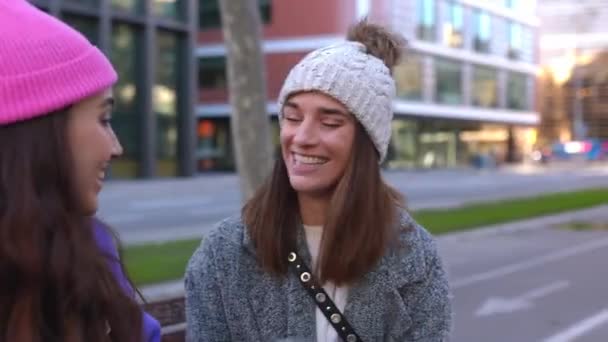 两个年轻而多样化的女人在城市街道上玩得很开心 千禧年的女性朋友 戴着冬帽 坐在长椅上享受都市背景的乐趣 友谊概念 — 图库视频影像