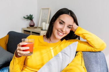 Genç güzel bir kadının gülümseyen portresi. Domates suyu içiyor. Evdeki kanepede dinleniyor. Sağlıklı yaşam tarzı ve refah kavramı.