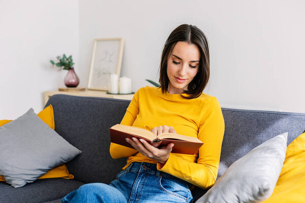 Relaxed young adult woman reading a book sitting on couch at her home.