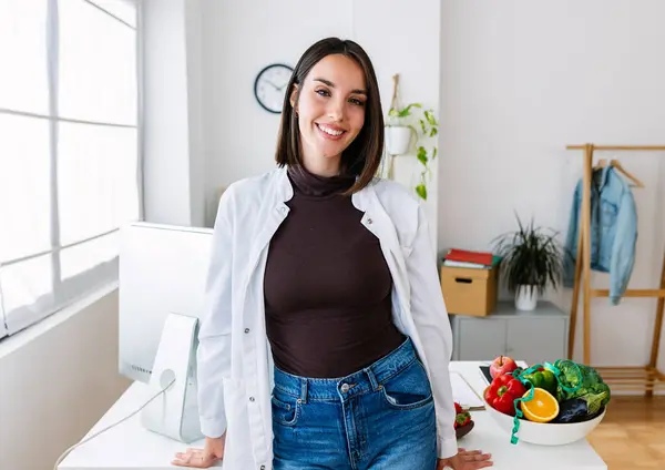 Smiling portrait of young beautiful female nutritionist doctor standing over office workplace. Hospital worker in white coat looking at camera at clinical desk consultation. Healthcare people concept