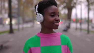 Kulaklıklı genç Afrikalı kadın şehir sokağında dikiliyor. Gülümseyen Afro-Amerikan kadın jenerasyonu..
