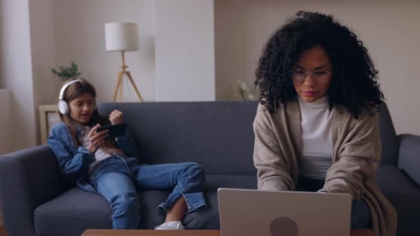 若い女性は スマートフォンでビデオゲームをする娘と自宅でソファーに座ってラップトップに取り組んでいます ファミリーコンクリレーション — ストック動画