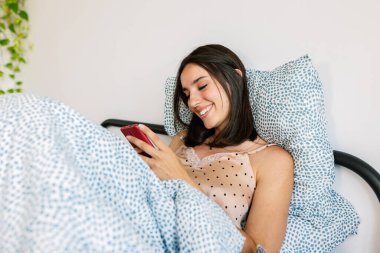 Genç, mutlu bir kadın, telefon uygulamasında yatarken sosyal medya içeriğini izlerken eğleniyor. Milenyum kadını uyandıktan sonra akıllı telefondan komik mesajlar okuyor..