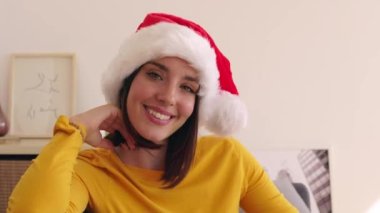 Noel Baba şapkalı güzel bir kadının portresi. Evdeki kanepede oturmuş kameraya gülümsüyor..