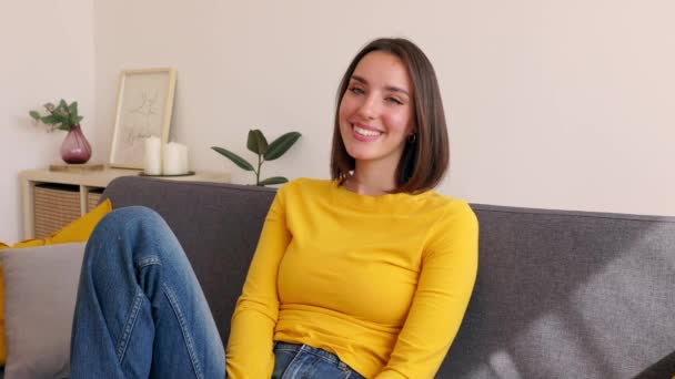 自宅でソファーに座ってカメラで微笑んでいる美しい若い大人の白人女性 — ストック動画