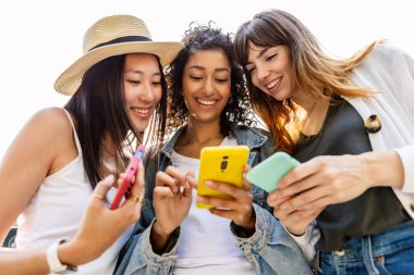 Dışarıda akıllı telefon kullanan mutlu bir grup genç kadın. Cep telefonuyla bağlanmış üç farklı bayan arkadaş sosyal medya içeriğini çevrimiçi izliyor. Teknoloji yaşam tarzı konsepti.