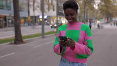 Şehir caddesinde cep telefonuyla yürüyen şık bir Afrikalı kadın. Teknoloji ve şehir yaşam tarzı konsepti.