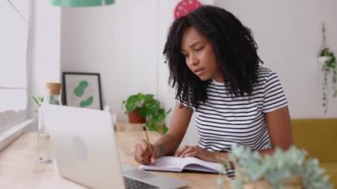 Evde dizüstü bilgisayar kullanırken not alan Brezilyalı genç bayan. Milenyum Latince kıvırcık kadın çevrimiçi sanal video dinlemeyi öğreniyor. İş ve eğitim kavramı.