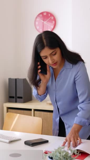 オフィスで携帯電話で話しているプロのインドのビジネスマン 成功した女性マネージャーは コンピュータ上のいくつかの情報をチェックしながら同僚に電話しました 縦のアスペクト ビデオ — ストック動画
