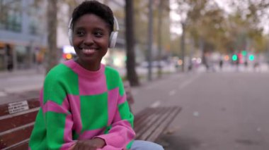 Kulaklıkla müzik dinleyen Afrikalı genç bir kadın şehir bankında oturuyor. Siyahi turist kadın dışarıda dinleniyor. Seyahat ve insan konsepti.