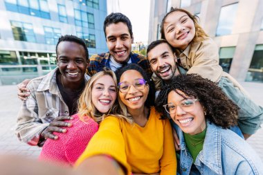 Çeşitli öğrenci grupları dışarıda cep telefonu uygulaması ile selfie çektirirken eğleniyor. Birleşik çok ırklı arkadaşlar gülerek ve şehir caddesinde eğlenerek. Arkadaşlık ve gençlik kavramı.