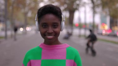 Kulaklıklı, sokakta kameraya bakan Afrikalı genç bir kadının portresi. Gülümseyen Afro-Amerikan kadın nesli z hipster kentsel arka planda gülümsüyor.
