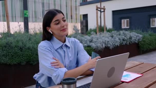 若いビジネス女性は 屋外でラップトップと座っている間 ワイヤレスイヤホンを使用して電話で話しています テクノロジーとビジネスコンセプト — ストック動画