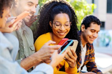 Milenyum öğrencileri kampüs kolejinde cep telefonlarını birlikte kullanıyorlar. Afrikalı Amerikalı bir kadın cep telefonu uygulamasında arkadaşlarıyla dışarıda otururken sosyal medya içeriğini izlerken eğleniyor..