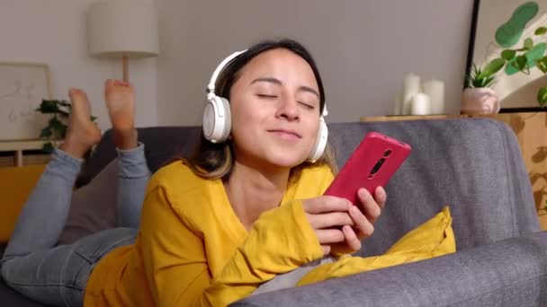 Kulaklıklı Mutlu Genç Bir Kadın Evdeki Koltukta Uzanan Akıllı Telefon — Stok video