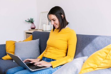Evdeki koltukta oturan dizüstü bilgisayarını kullanan neşeli bir milenyum kadını. Teknoloji ve e-öğrenme kavramı.