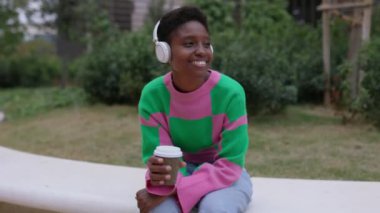Genç Afrikalı bir kadın kablosuz kulaklıkla müzik dinlerken şehir parkında oturuyor. Milenyum siyahi dişisi şehir molasının tadını çıkarıyor..
