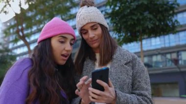 İki yetişkin kadın cep telefonuna bakıp sosyal medya haberlerinin şehir caddesinde yan yana durduğunu tartışıyor. Teknoloji ve medya kavramı.