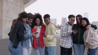 Dışarıda cep telefonuyla gülen bir grup mutlu insan. Gülümseyen çok ırklı öğrenci arkadaşlar akıllı telefon uygulamasında komik sosyal medya videoları izlerken eğleniyorlar. Gençlik toplumu kavramı