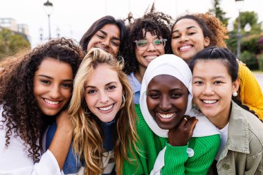 United portrait of young multiracial girls smiling at camera standing together outdoors. Millennial female friends feeling hugging each other smiling and posing for a photo. Women community concept. clipart