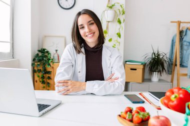 Portrait of young beautiful nutritionist woman smiling at camera at medical consultation. Professional healthcare worker concept clipart