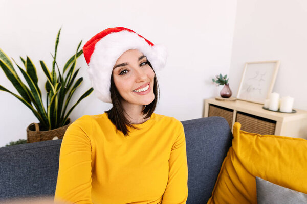 Pretty millennial woman in Santa Claus hat taking selfie portrait at christmas holidays sitting on sofa at home