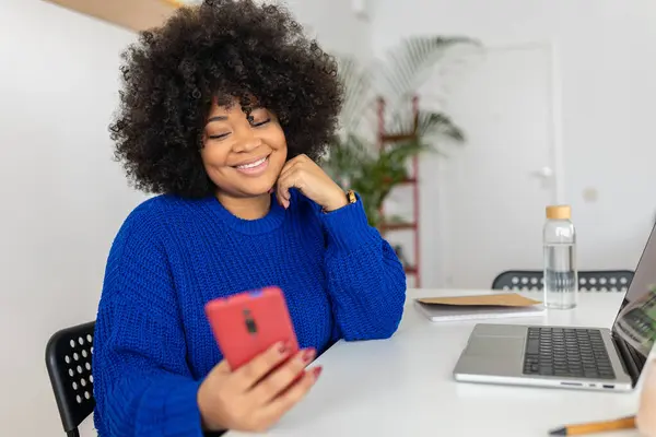 Happy young african woman with laptop using mobile phone sitting on table at home. Happy black female looking at smart phone screen.