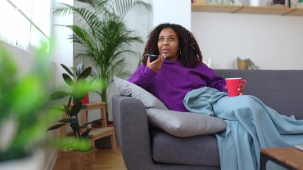 モバイルアプリを使用している若いアフリカの若い女性は ソファーで休んでいる間 オーディオボイスメッセージを送信し 冬の季節にホットドリンクを飲みます 自宅で快適な朝のコンセプト — ストック動画