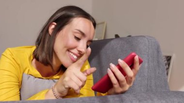 Evde cep telefonu kullanan gülümseyen bir kadın. Genç ve güzel bir kadın cep telefonu ekranına bakıyor, kanepeye uzanmış uygulamanın sosyal medya içeriğinin tadını çıkarıyor. Teknoloji konsepti.