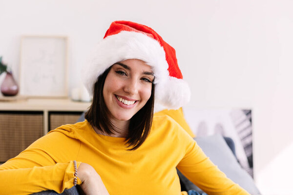 Portrait of young pretty woman in Santa Claus hat smile at camera sitting on sofa at home during christmas vacation.
