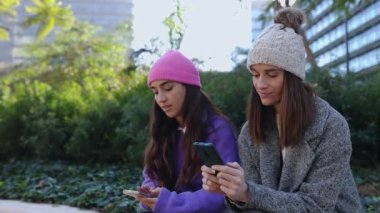 Dışarıda oturmuş akıllı telefon ekranına bakan iki genç ve rahat kadın. Kadın arkadaşlar kış günü şehir caddesinde sosyal ağlarda içeriği izlerken eğleniyorlar..