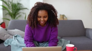 Kışın evdeki oturma odasında kanepede otururken battaniyeye sarılmış genç siyahi bir kadın dizüstü bilgisayar kullanıyor..
