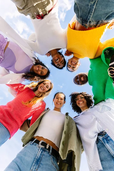 Low angle view vertical photo of young diverse women standing together in circle hugging each other and smiling at camera outdoors