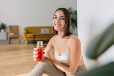 Happy young attractive woman smiling at camera holding bottle of tomato juice. Millennial fit female enjoying healthy drink after workout fitness routine at home. Sportive people and vegan meal. clipart